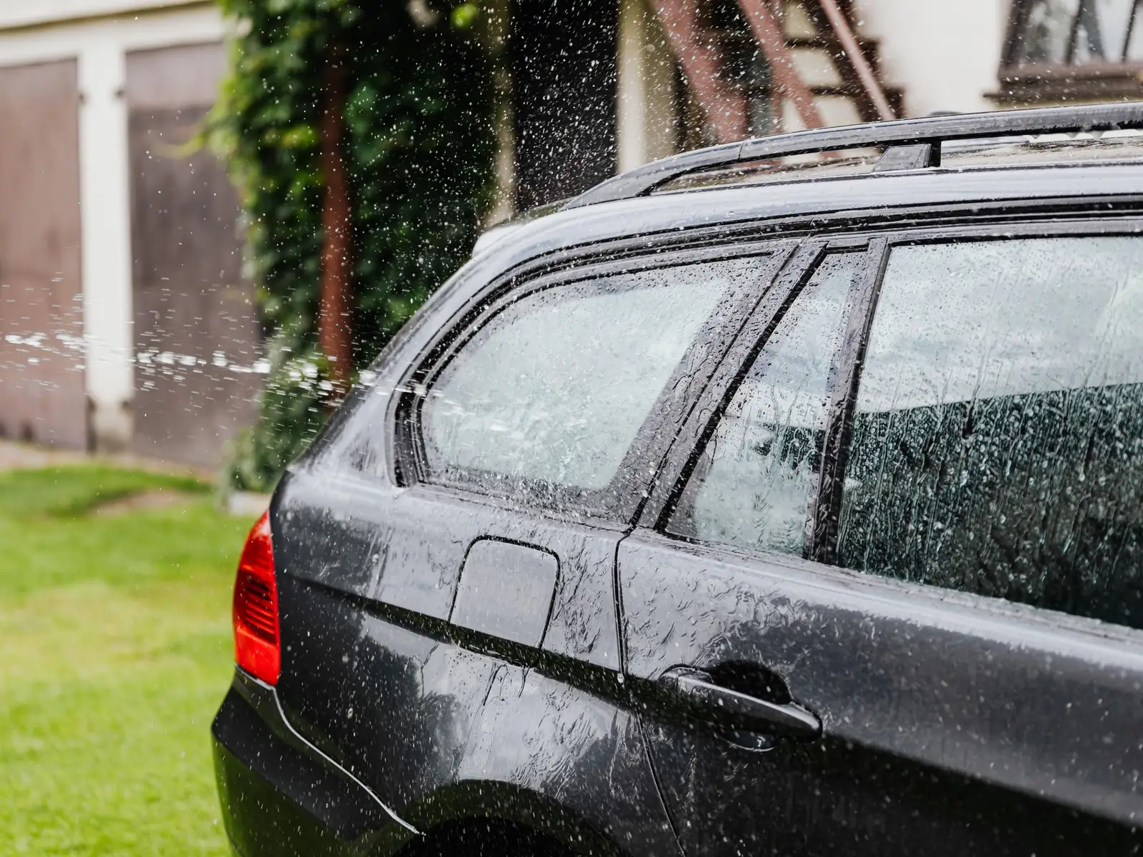 A car being washed by hand outside a house, water running down the paintwork and glass